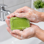 A person's hands covered in lathered soap wash are holding a green rectangular bar of Gyanveda soap over a white sink with a chrome faucet in the background.