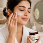 A woman in a white bathrobe applies cream to her cheek while holding a jar of Gyani Anti Aging Cream in a softly lit bathroom setting.