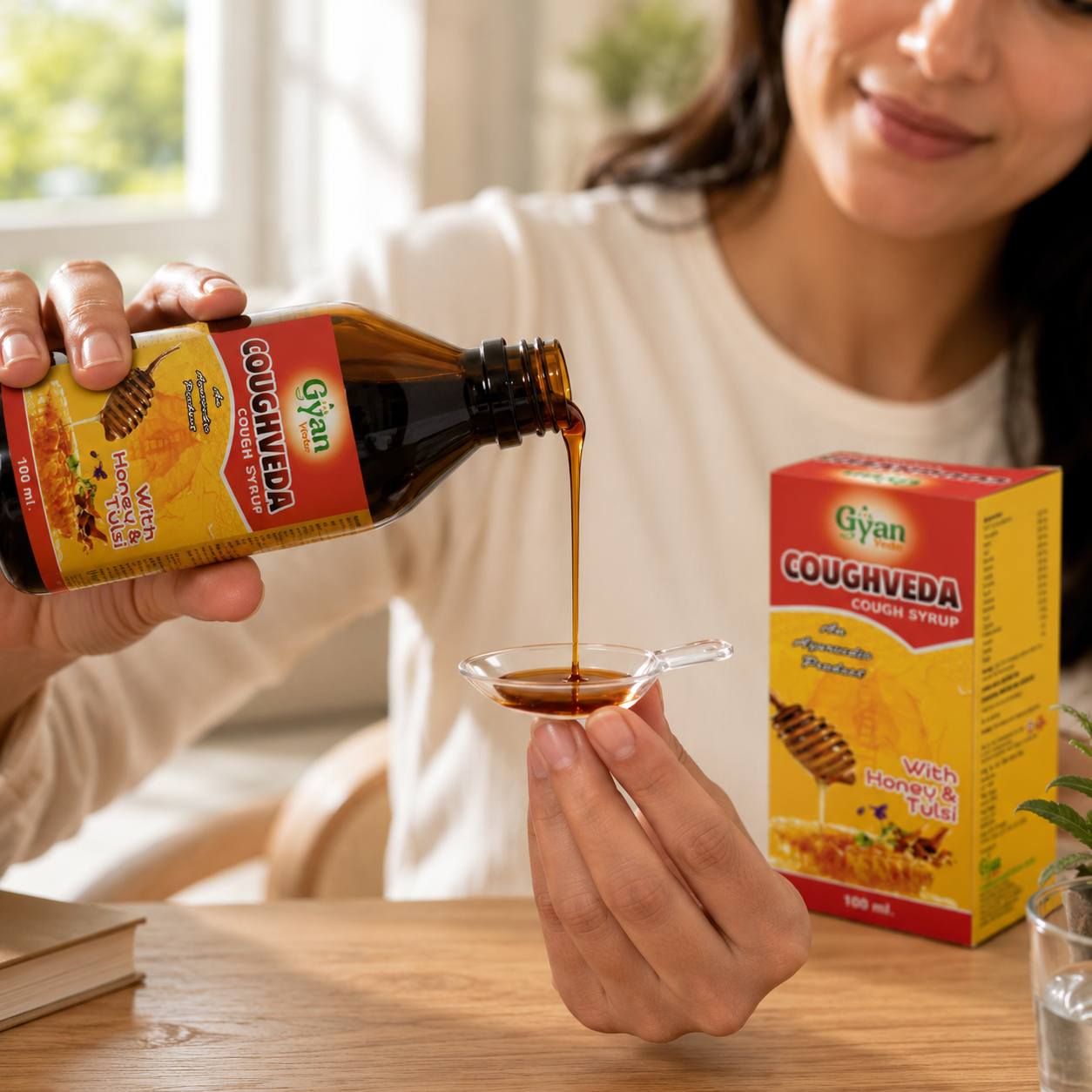 A woman pours dark brown cough med from a bottle labeled Gyan Coughveda into a small glass spoon, with the product box standing on a wooden table nearby.