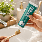 A hand holds a tube of Gyan Veda Toothpaste above a bamboo toothbrush, with toothpaste being squeezed onto the bristles in a bright bathroom setting.