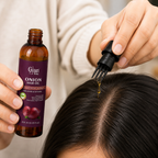 A close-up of a person applying onion hair oil from a small amber bottle with a dropper onto the center part of their dark hair. The person is wearing a cream-colored sweater, and the bottle label reads 'Gyan Vida Onion Hair Oil'.