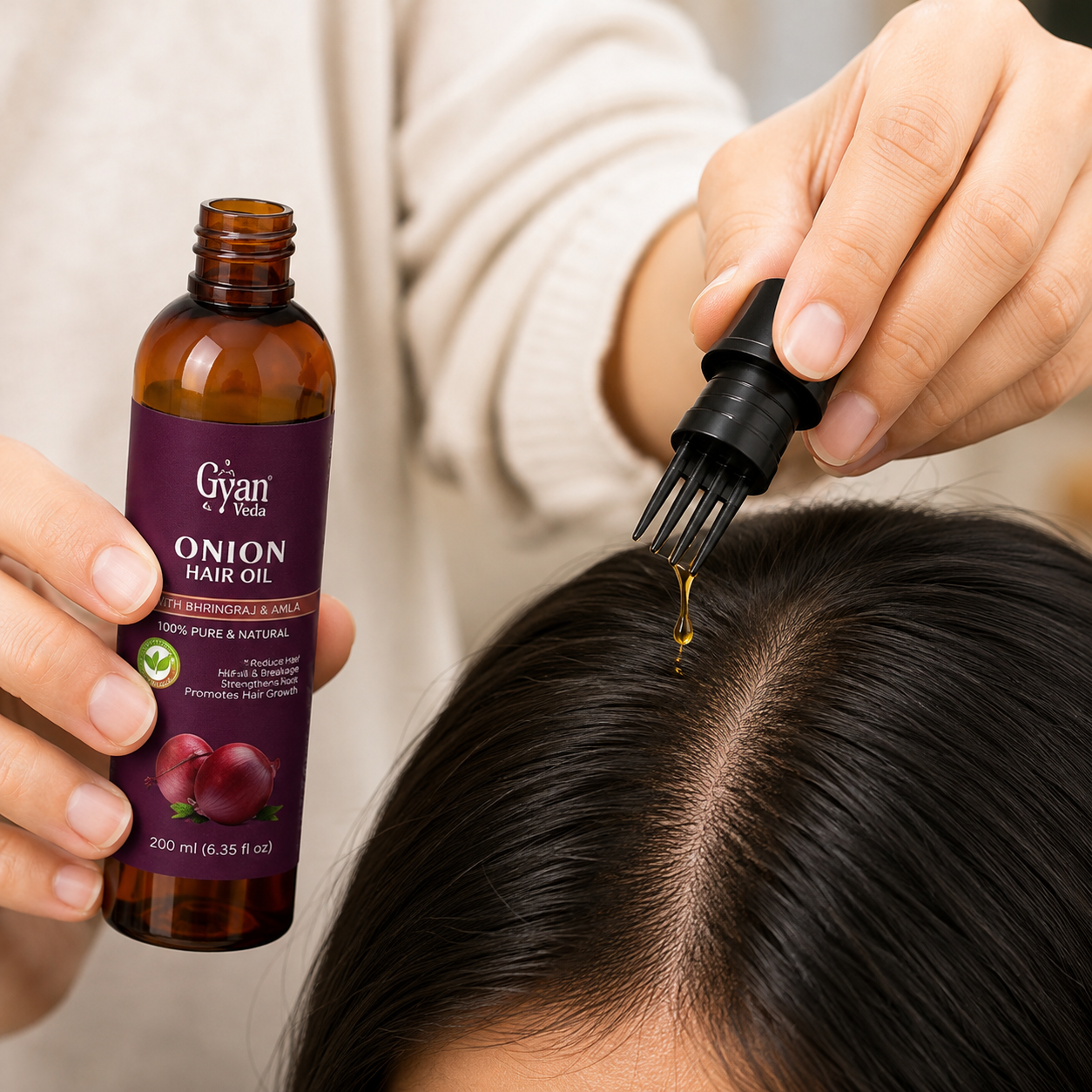 A close-up of a person applying onion hair oil from a small amber bottle with a dropper onto the center part of their dark hair. The person is wearing a cream-colored sweater, and the bottle label reads 'Gyan Vida Onion Hair Oil'.