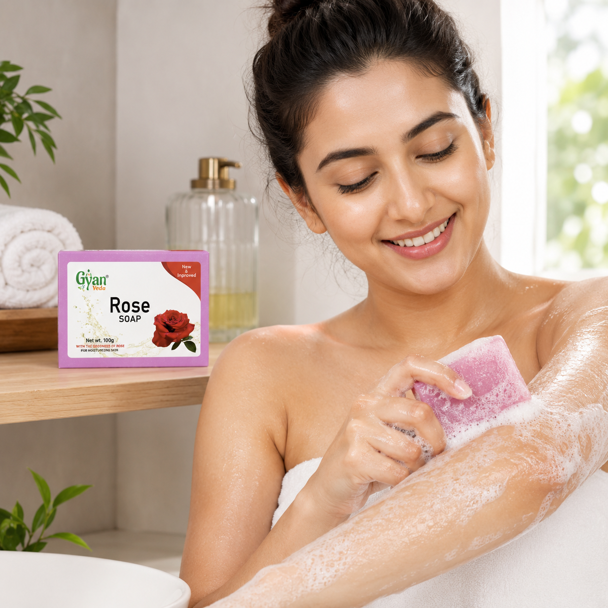 A woman wrapped in a white towel applies a pink rose-shaped soap bar to her arm in a bright bathroom setting. The product packaging, labeled 'gyanveda Rose Soap', is shown on a wooden shelf in the background.