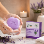 A person holds a lavender-colored bar of soap covered in white lather, with a purple and white Gyan Veda Lavender Soap box beside it on a marble surface. Lavender sprigs, a lit candle, and a soap dispenser are softly blurred in the background.